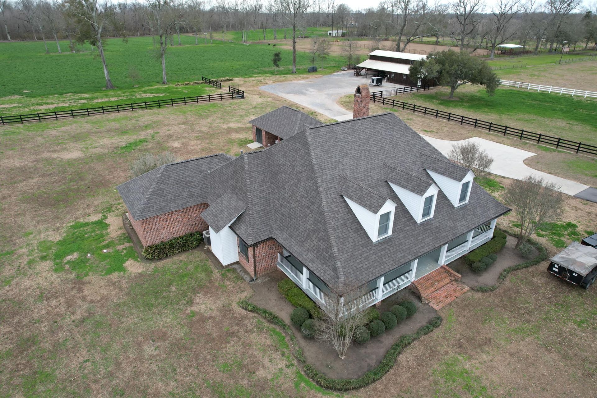 Aerial view of a large gray-roofed house with dormers on a rural property with fields and a driveway.
