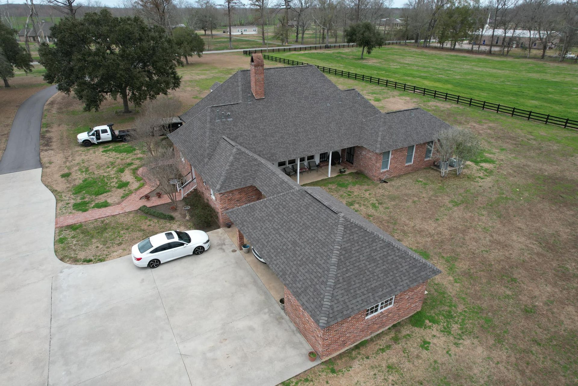 Aerial view of a brick house with a dark shingled roof, driveway, and two parked cars near a rural road.