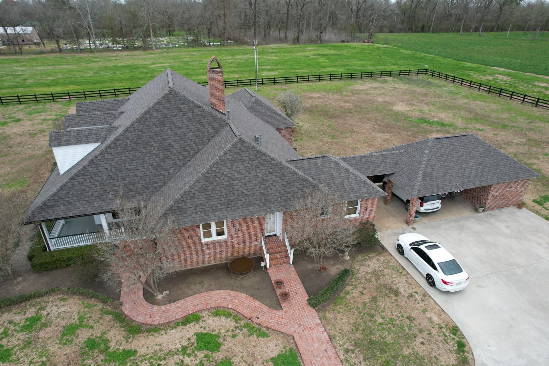 Aerial view of a brick house with a dark roof, driveway, white car, and surrounding green fields.