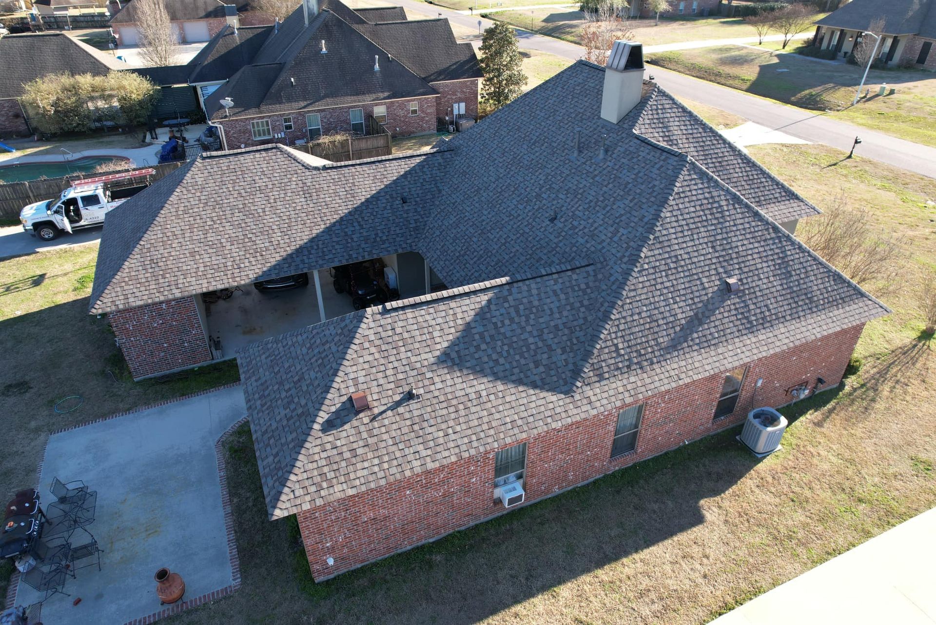Aerial view of a brick house with a dark shingled roof and driveway in a suburban neighborhood