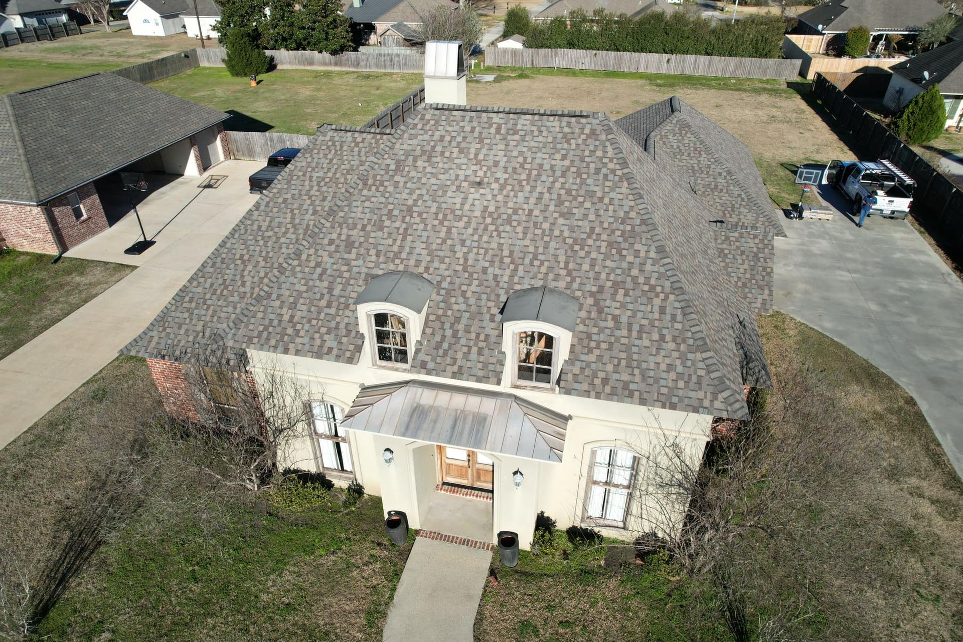 Aerial view of a beige house with a gray shingled roof and small front yard in a suburban neighborhood