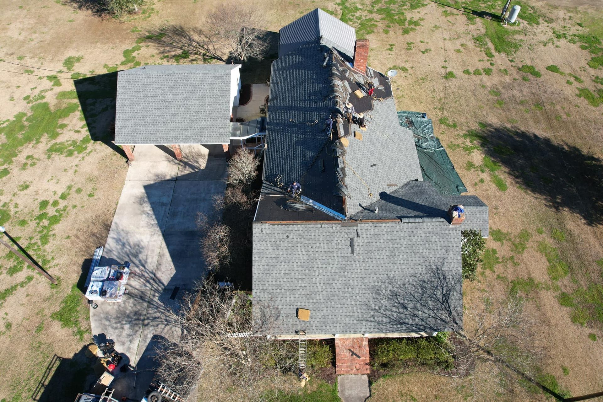 Aerial view of a house with gray roofs, a central dark roof, and surrounding bare yard.