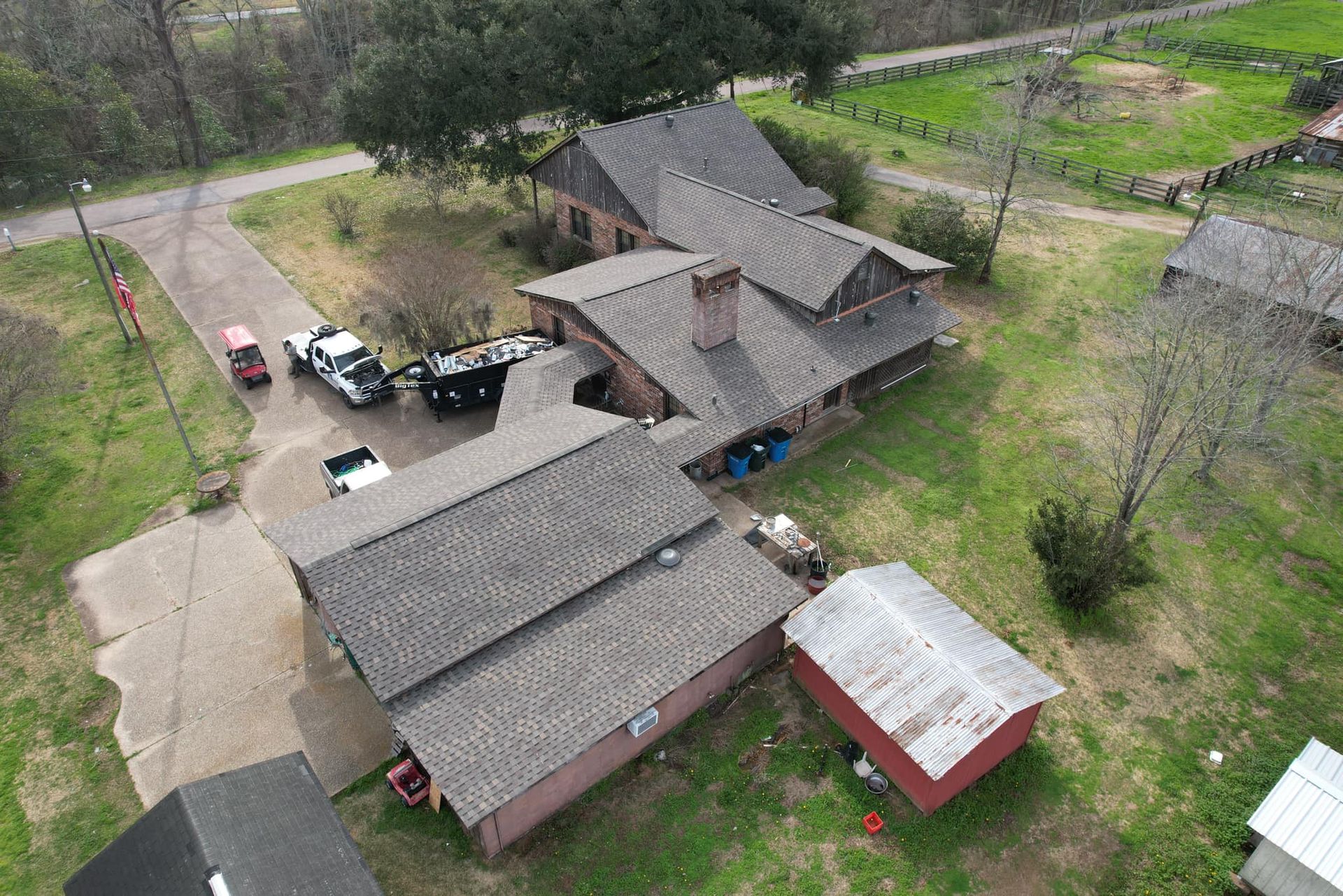 Aerial view of a rustic house with gray roofs, a red shed, and parked cars in a grassy yard
