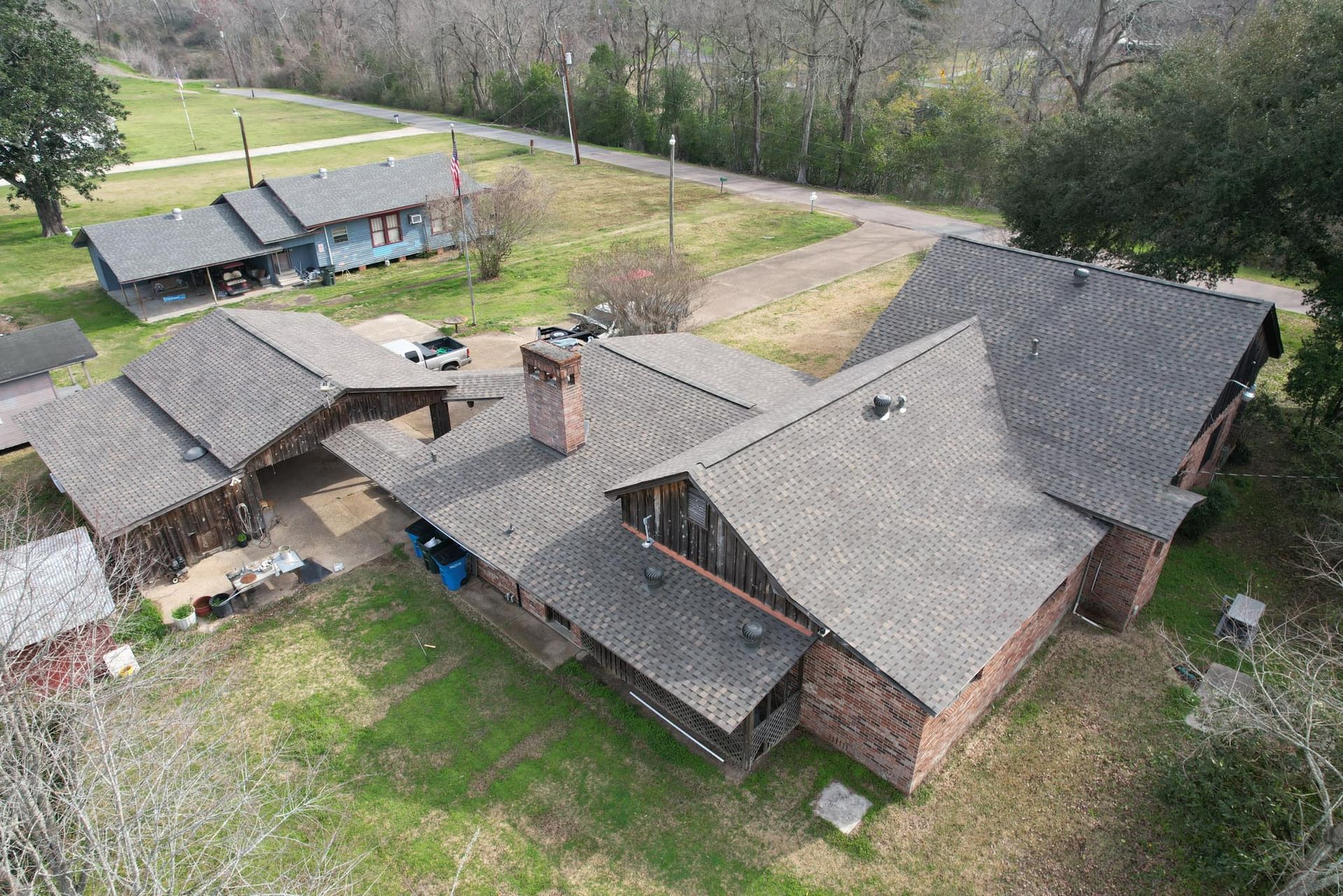 Aerial view of a brick house with gray roof, porch, and detached shed beside a road and trees.