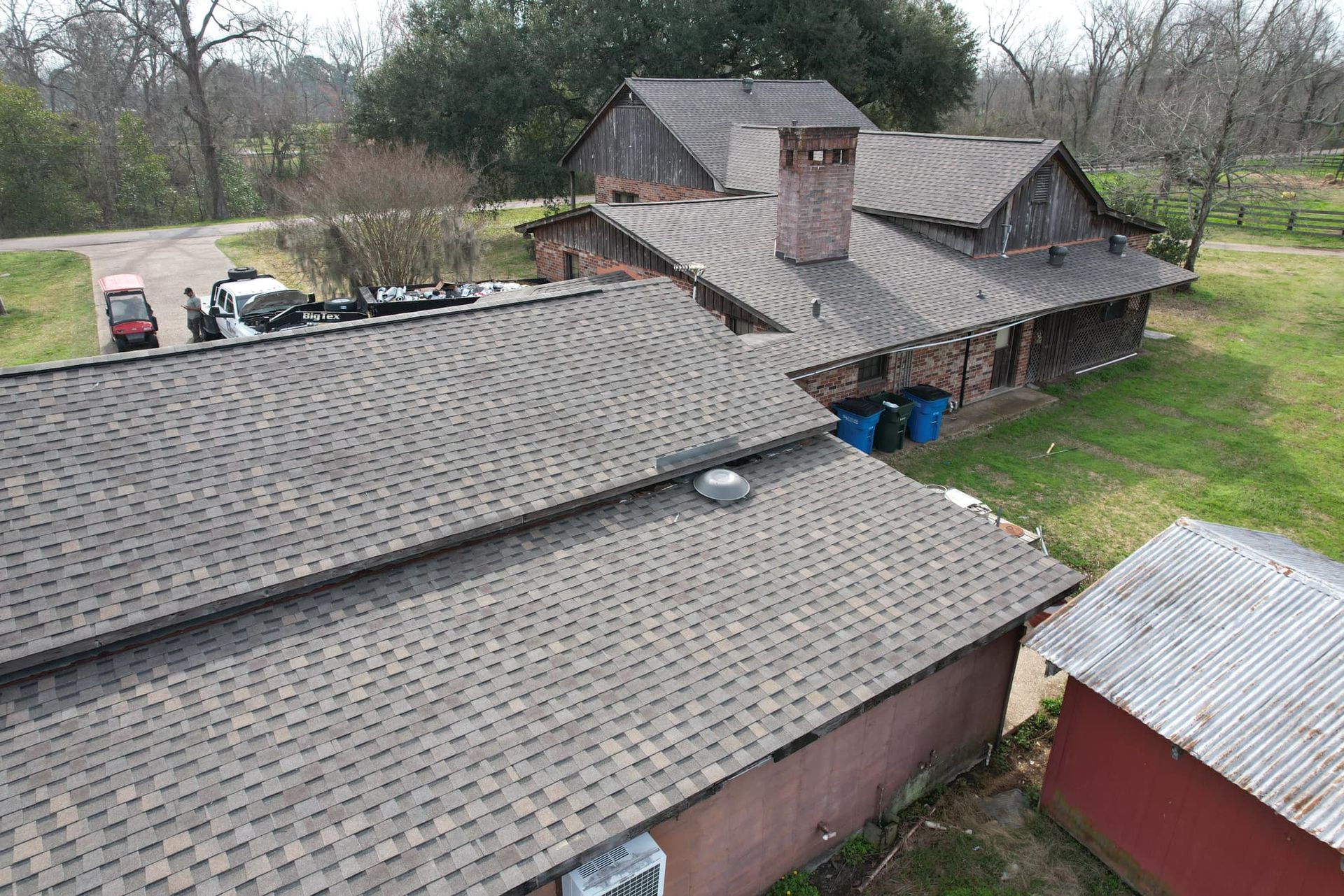 Aerial view of a rural farmhouse with gray shingle roofs, a brick chimney, and surrounding trees and grass.