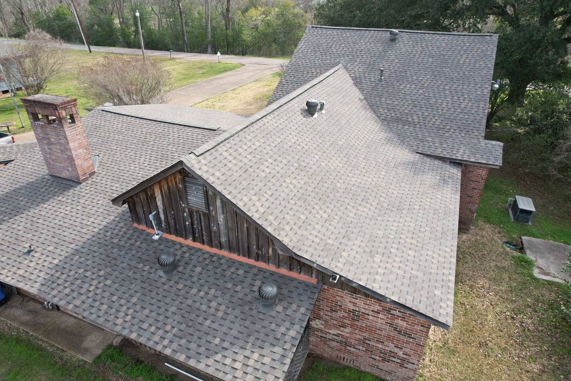 Aerial view of a house with a gray shingle roof, brick chimney, and wooded yard.