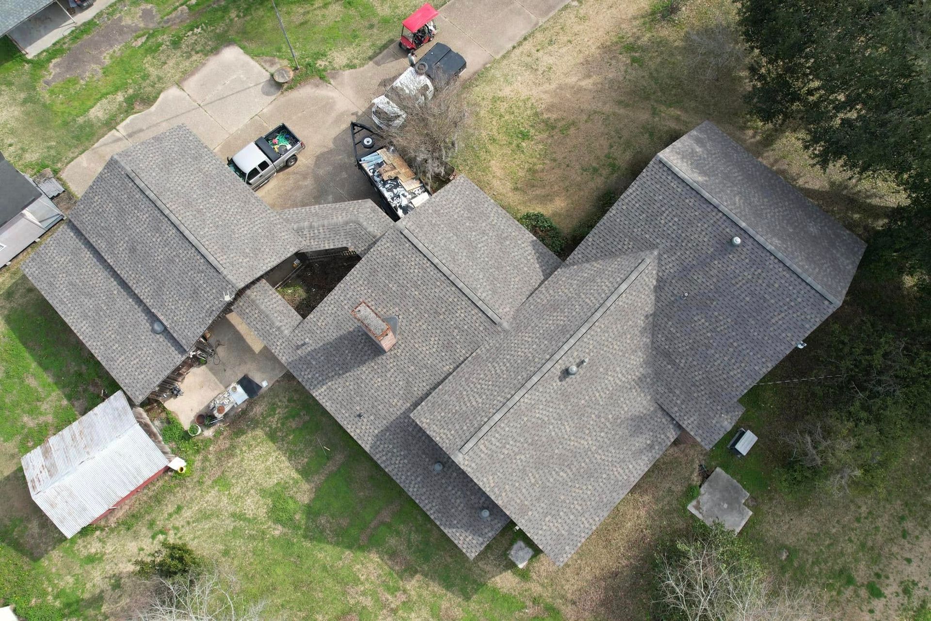 Aerial view of gray-roofed buildings and parked vehicles in a rural yard with grass and trees