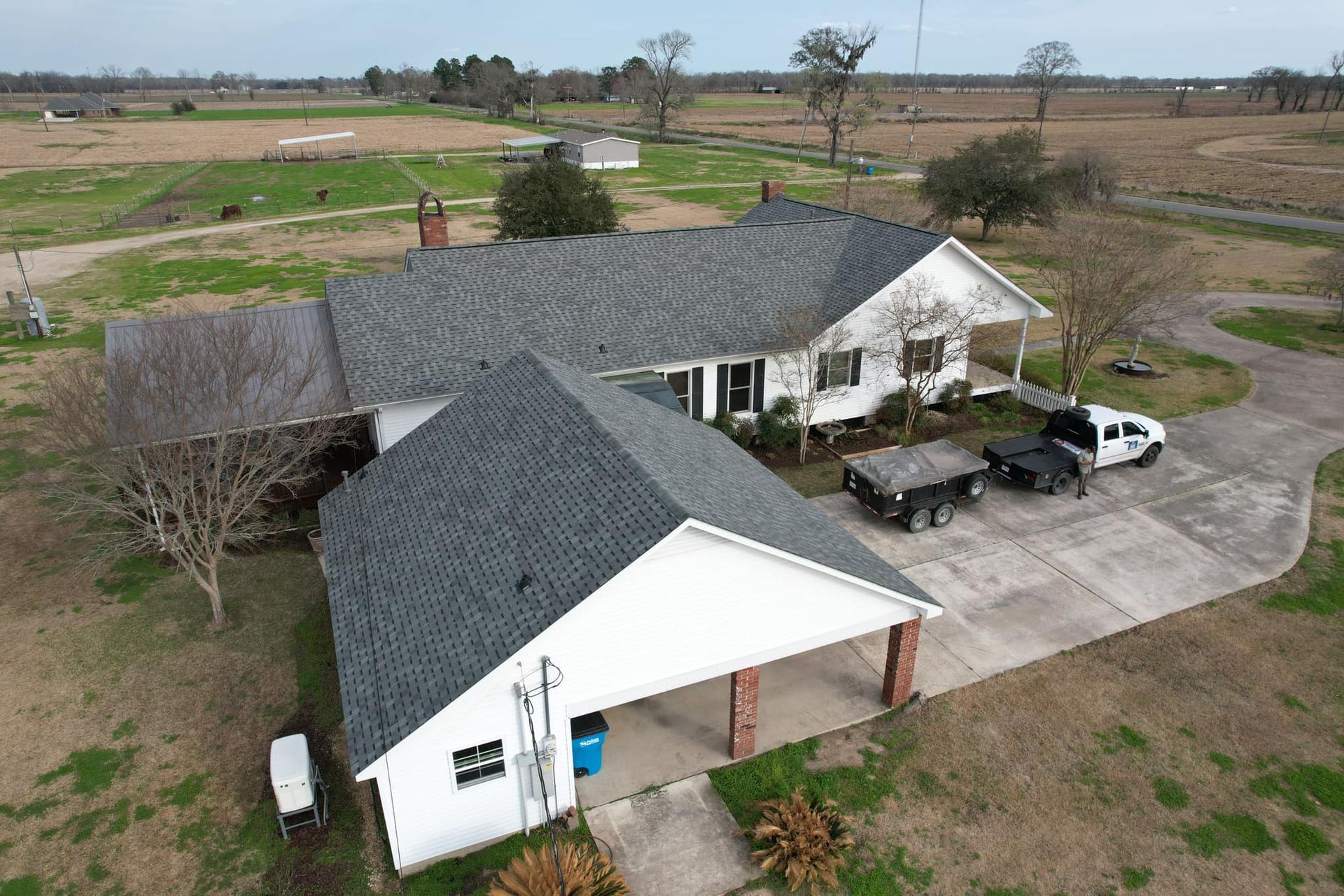 Aerial view of a white house with a gray roof, attached garage, driveway, and parked vehicles in a rural field.