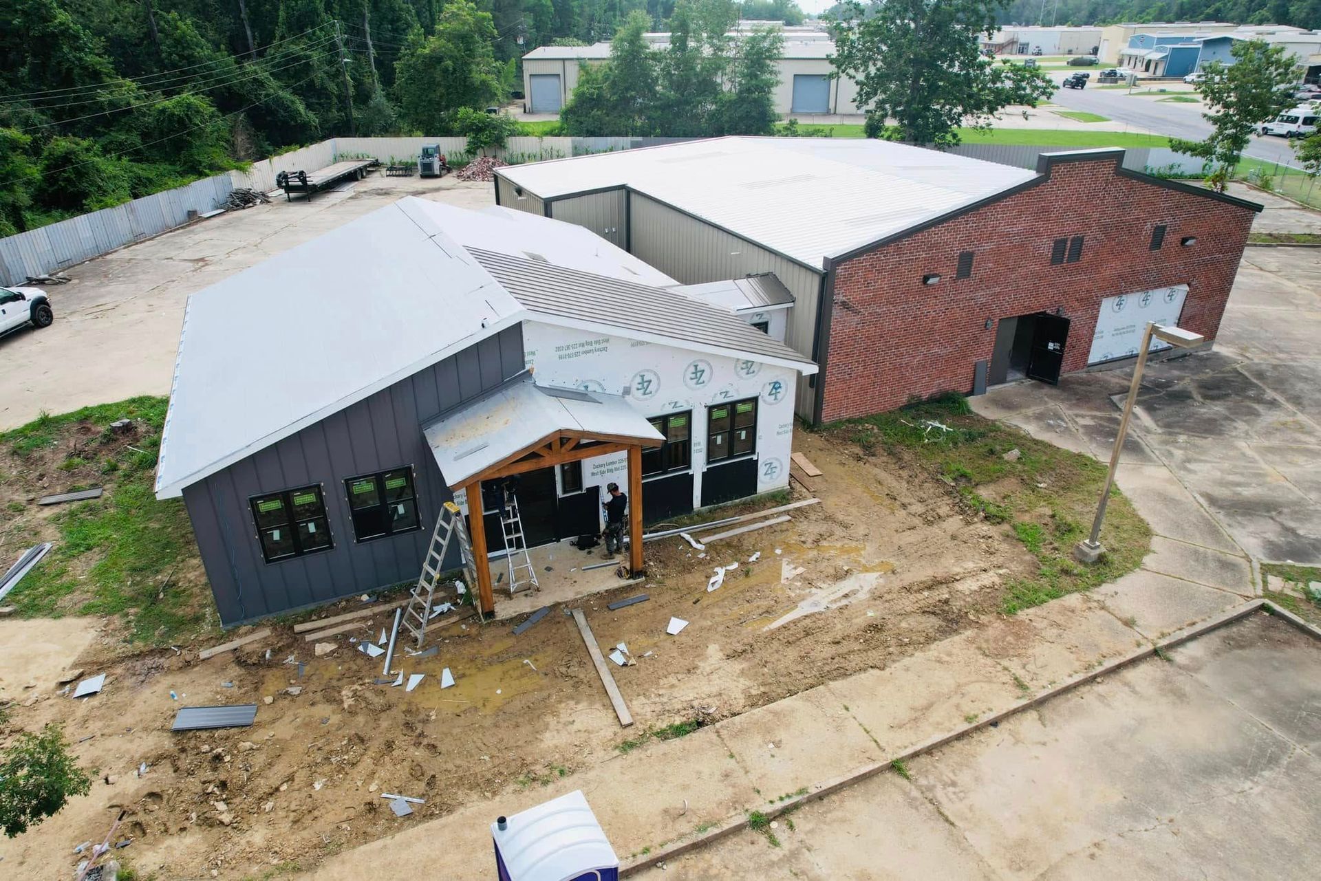 Aerial view of two connected buildings, one gray and one red brick, amid a dirt lot and trees.
