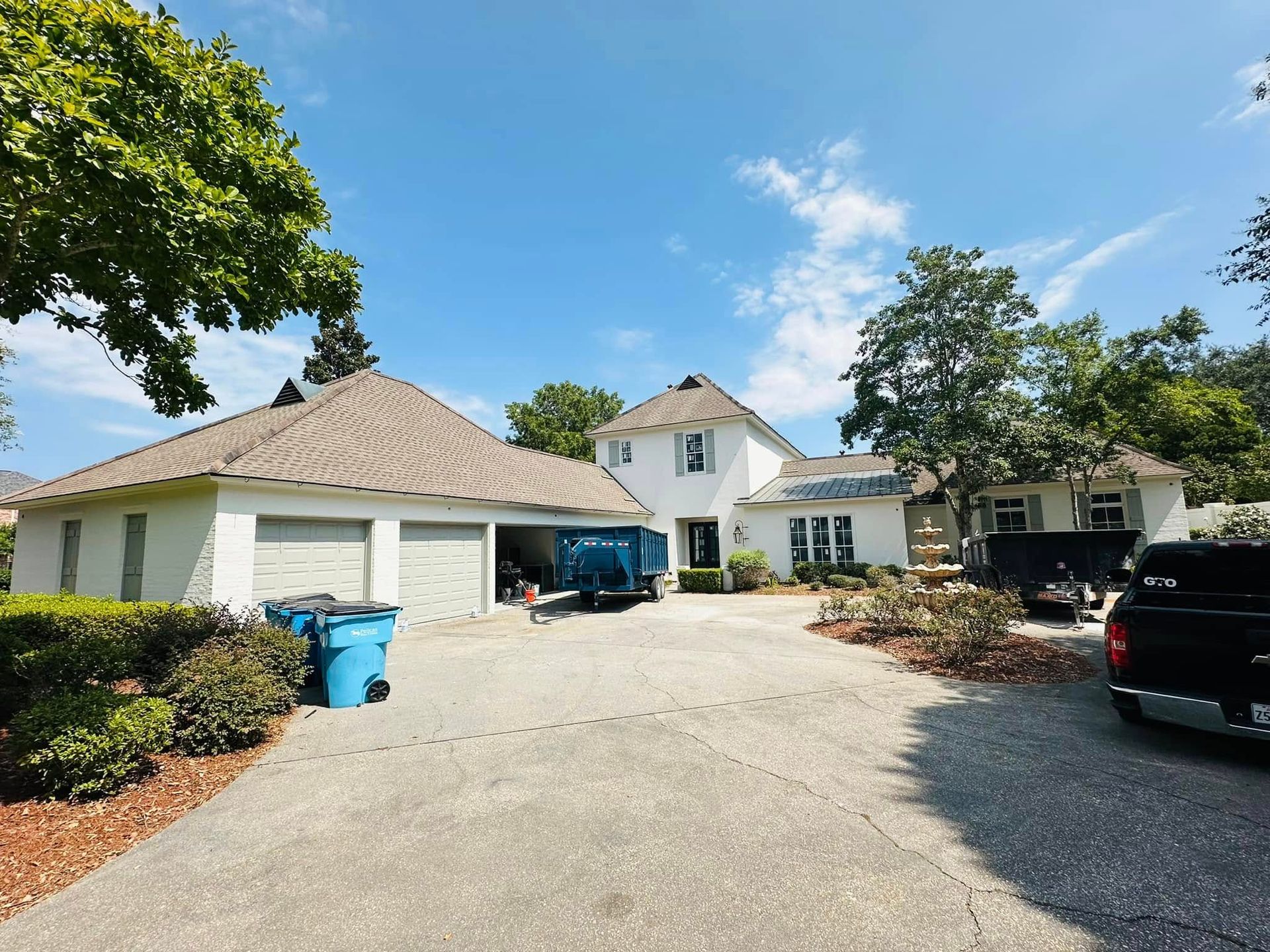 Driveway view of a white house and detached garage with blue trash bins and parked cars under a sunny sky