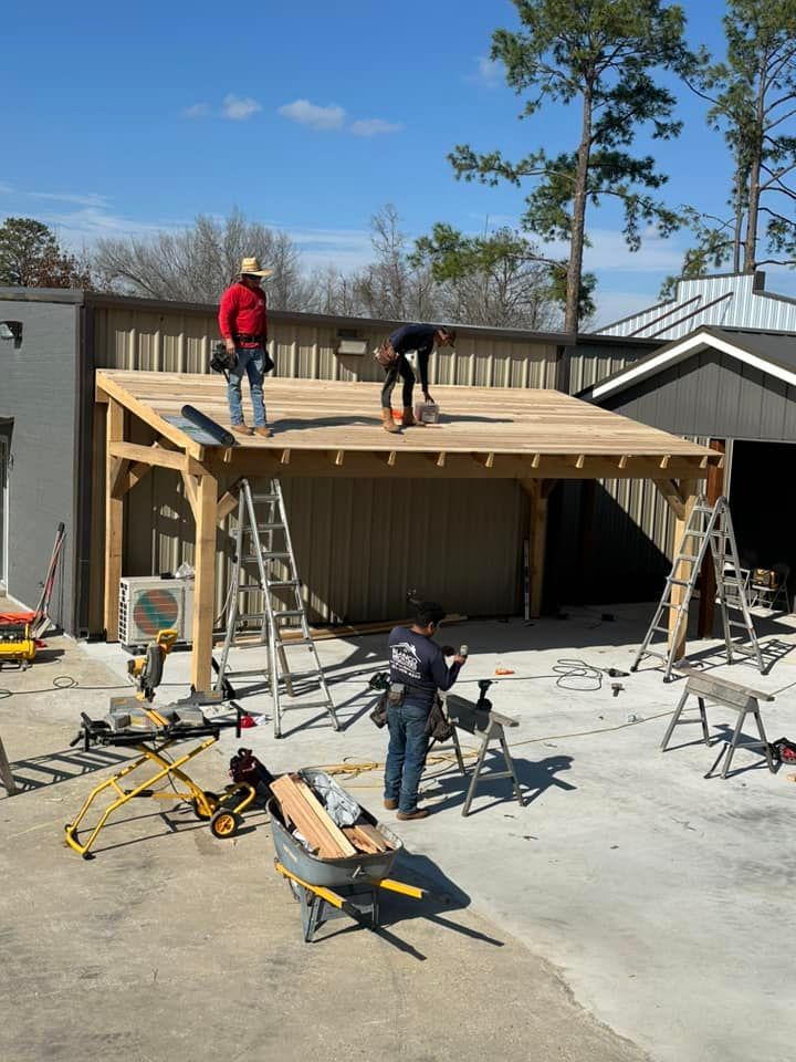 Workers building a wooden roof frame over a driveway, with ladders and tools on a sunny day