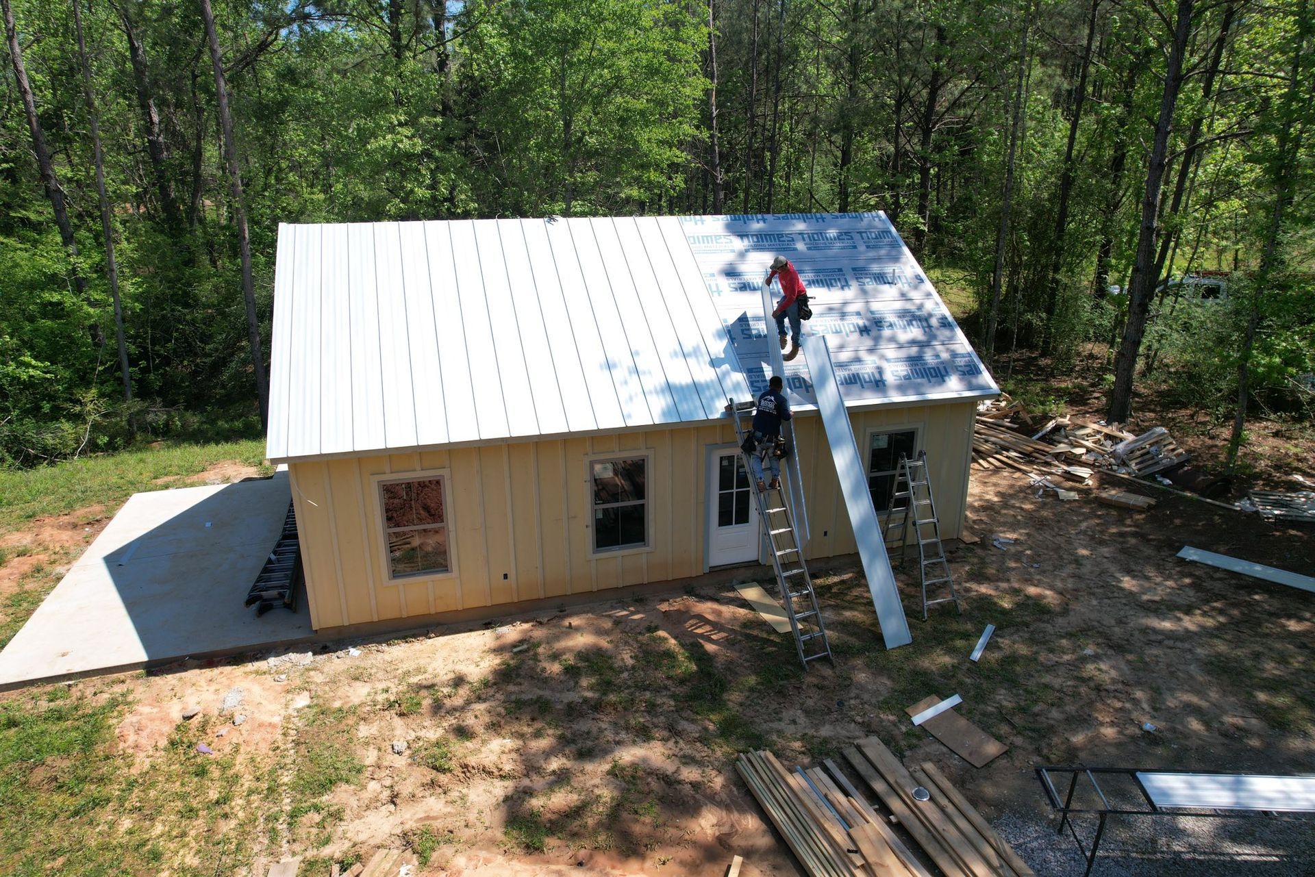 Worker on a white roof installing panels on a tan house in a wooded area, with a ladder against the roof.