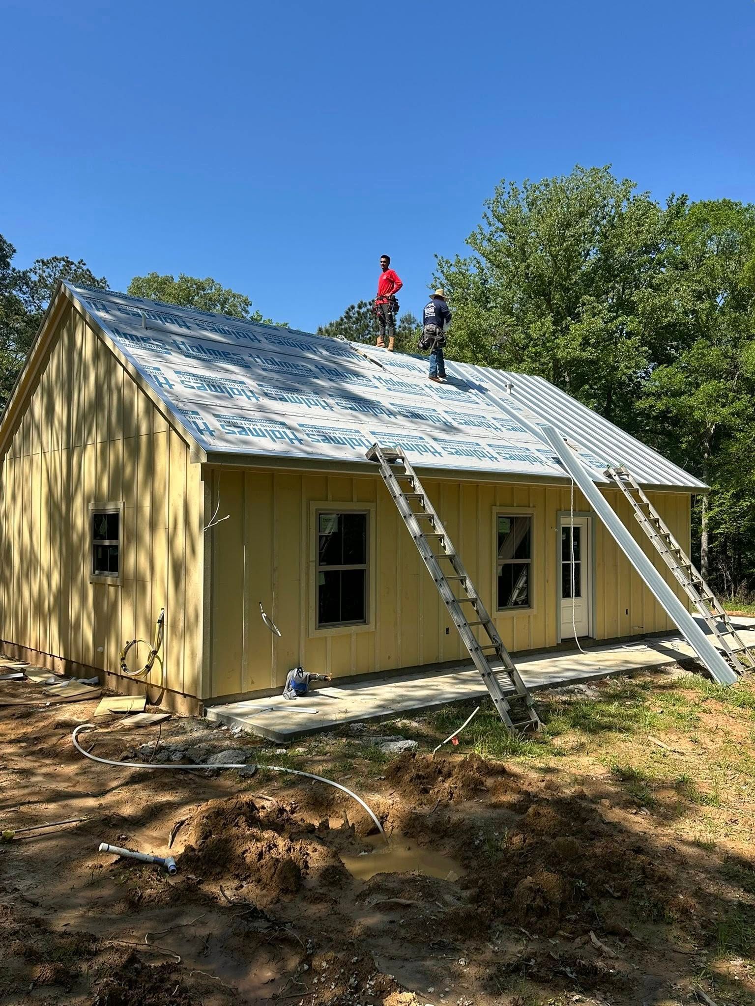 Two workers on a ladder reroofing a small yellow house with a silver metal roof under a blue sky