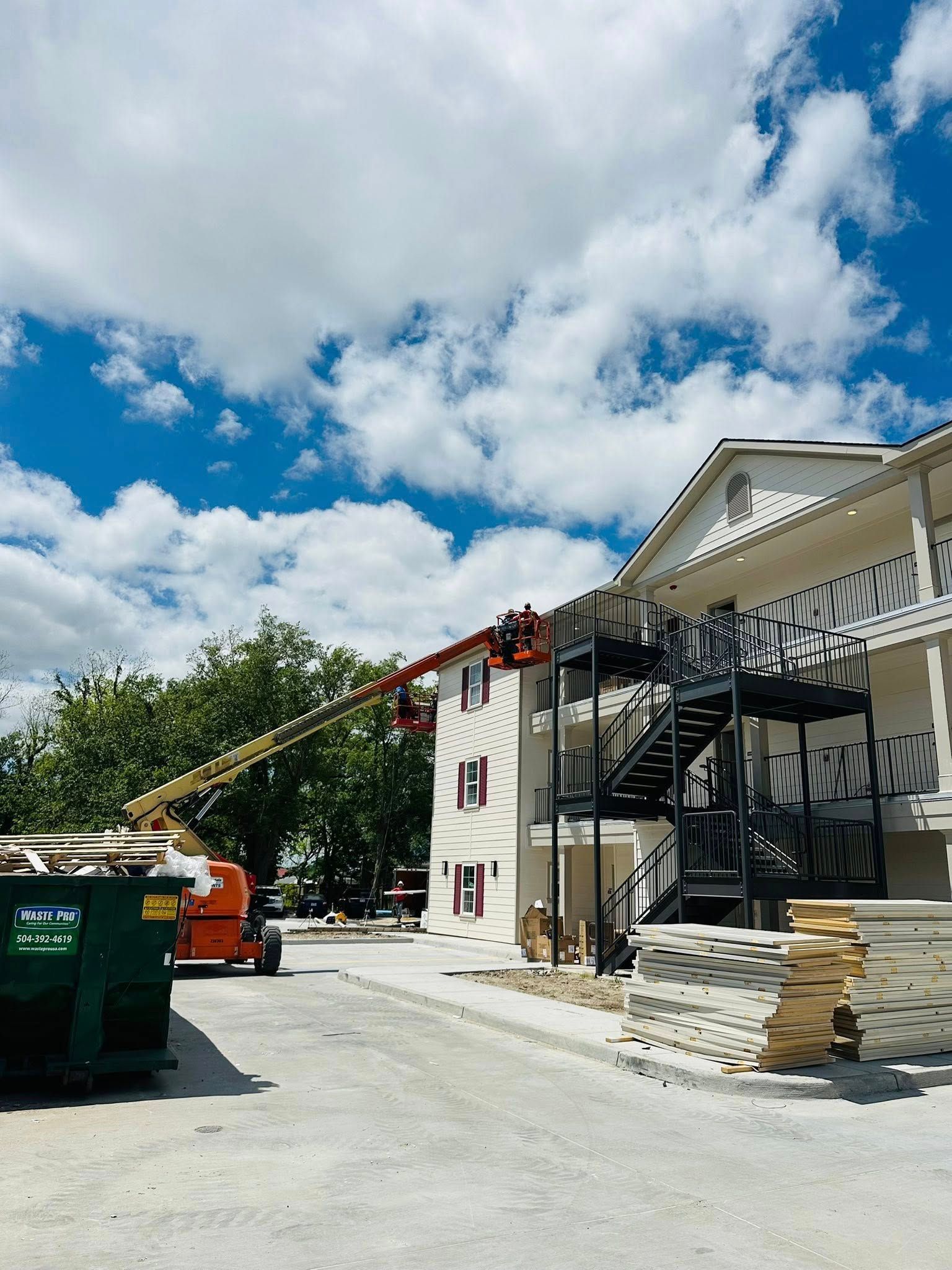 Construction site with white building, exterior stairs, red crane, and dumpsters under a partly cloudy sky