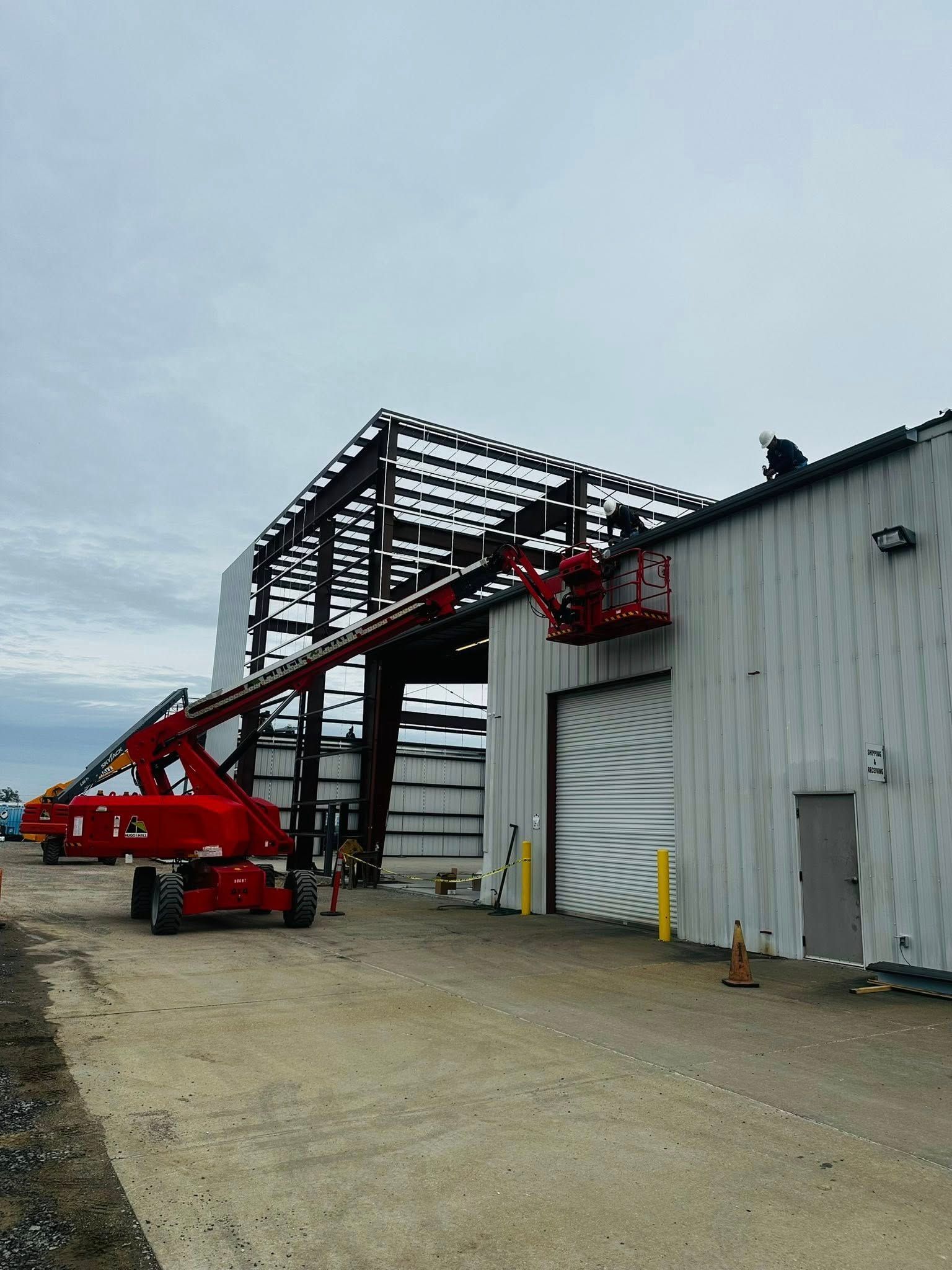 Red boom lift raised beside a metal industrial building under construction on a gravel lot.