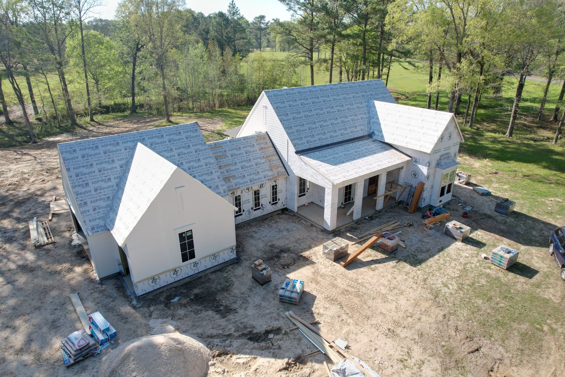 Aerial view of a new house under construction with gray roofing, surrounded by trees and dirt work.