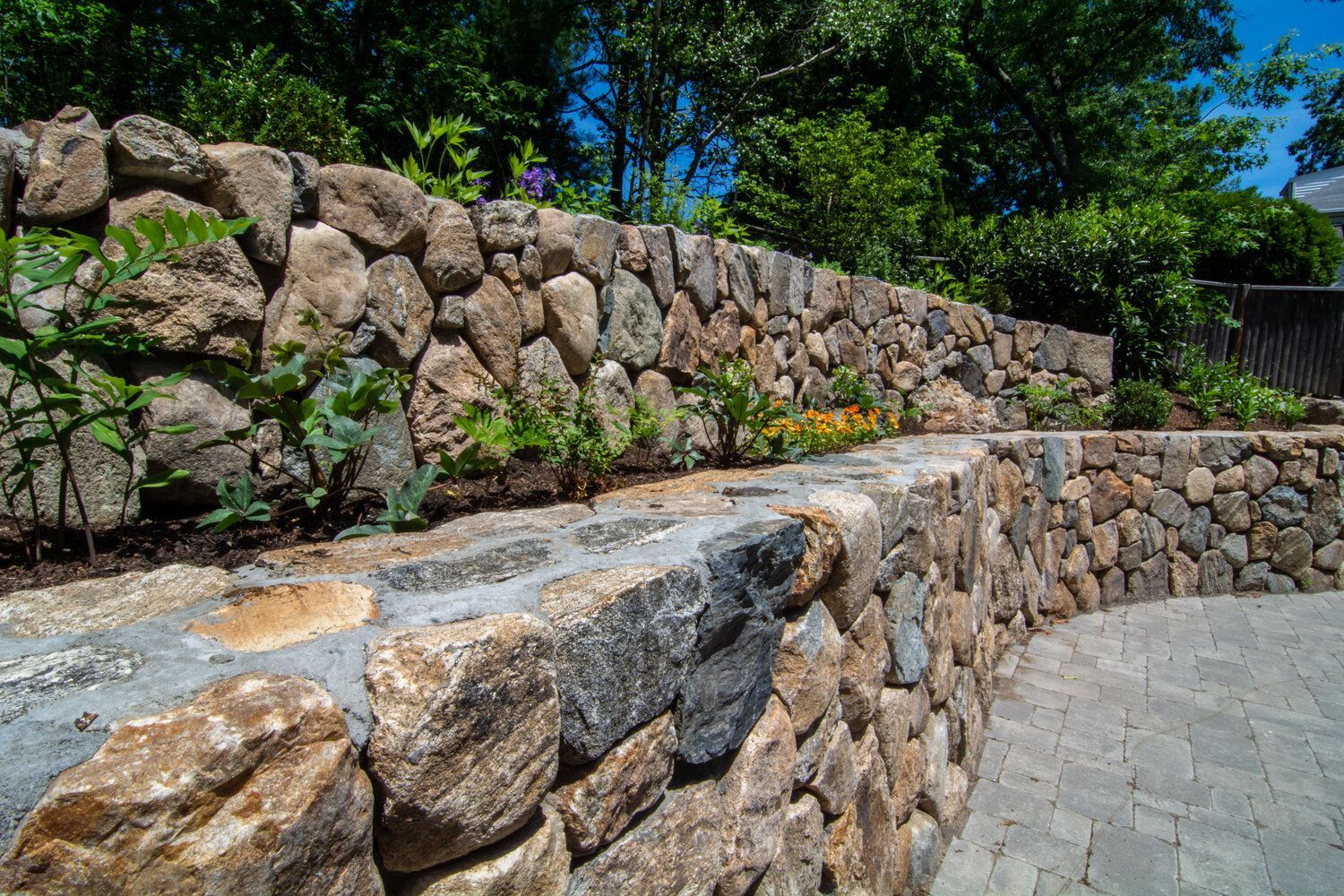 A stone wall surrounds a brick walkway in a garden.