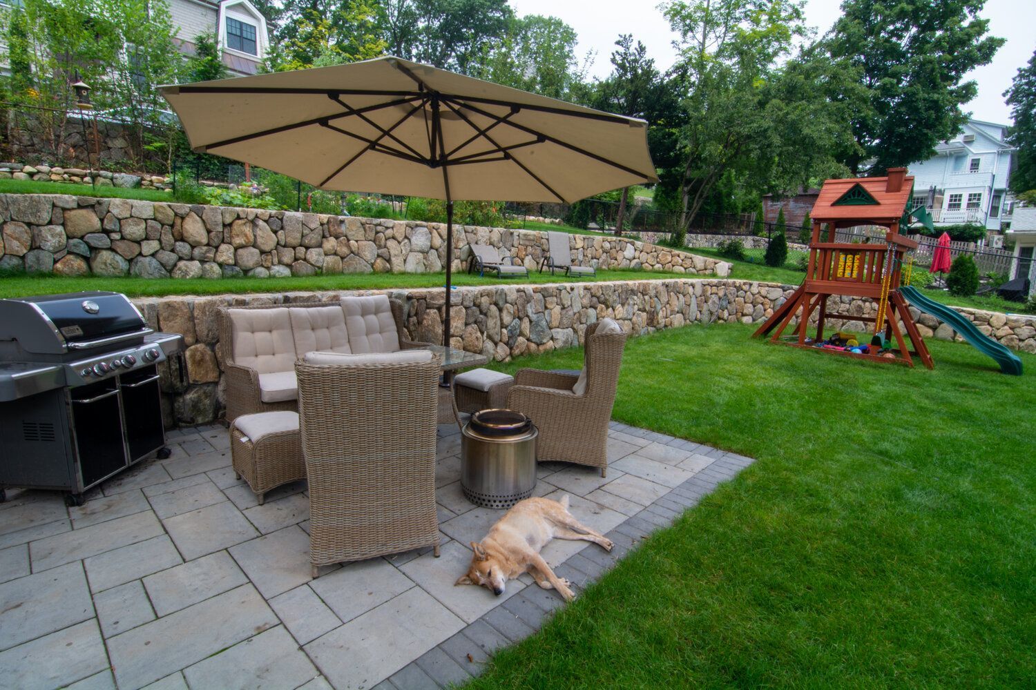 A dog is laying under an umbrella on a patio in a backyard.