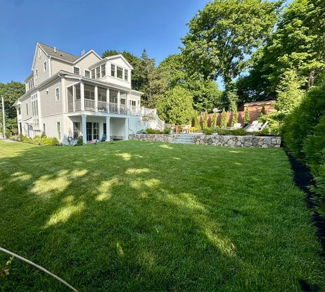 Large white house with wraparound porch beside a lush green lawn and trees on a sunny day