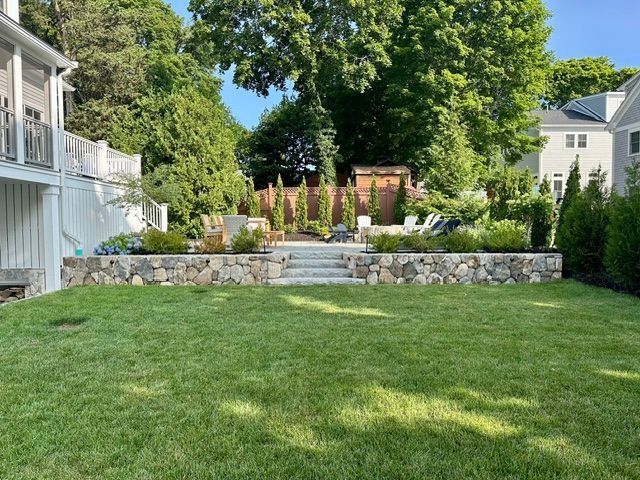 Landscaped backyard with stone retaining wall, steps, flowerbeds, and green lawn beside a white house