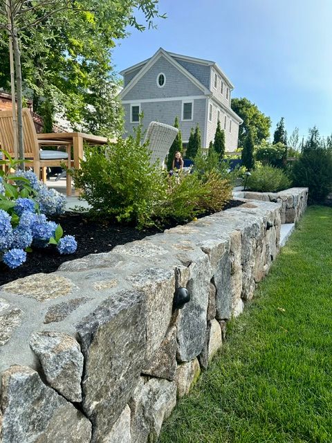 Stone retaining wall with flowers beside a green lawn, leading to a gray house on a sunny day