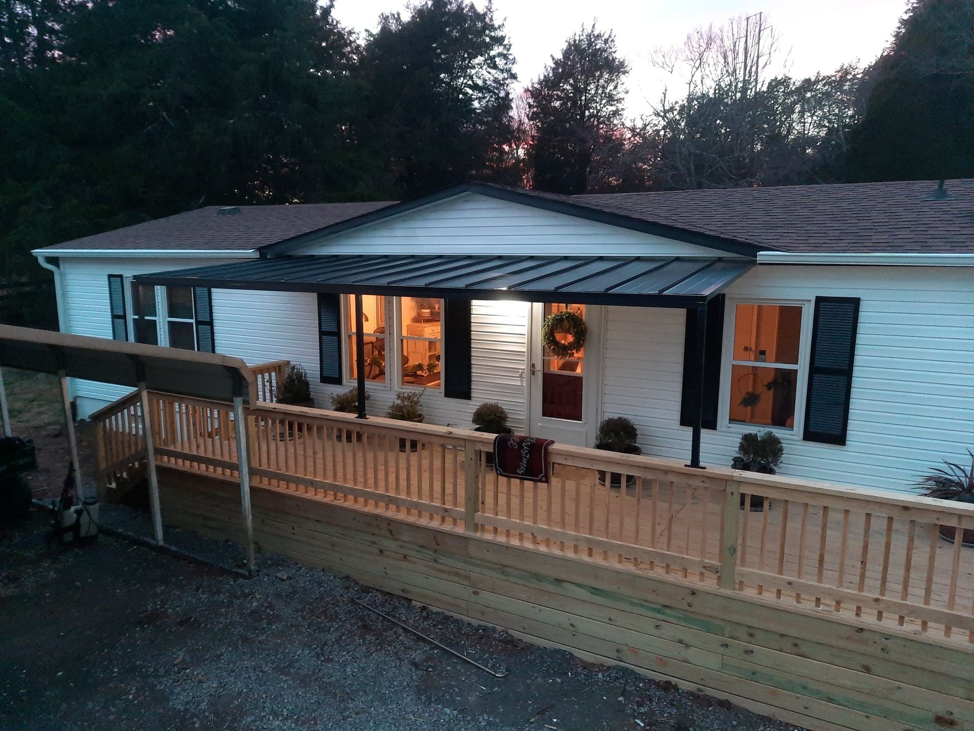 A manufactured home with white siding, a new wooden deck with railings, and a black metal porch awning at dusk.