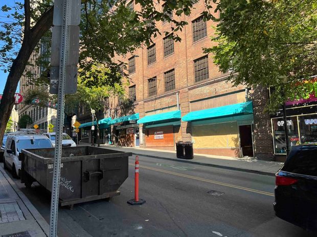 A street-level view of a brick building with turquoise awnings, a large dumpster, and parked vehicles on a city street.