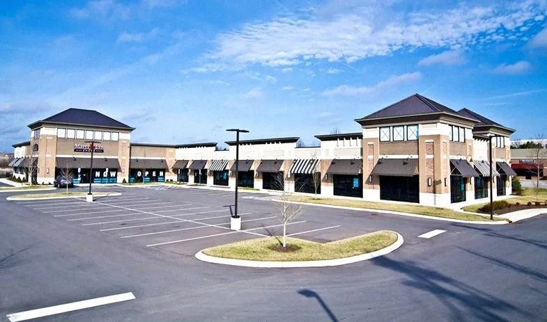 A modern, U-shaped retail strip center with tan brick facades, dark awnings, and an empty parking lot under a blue sky.