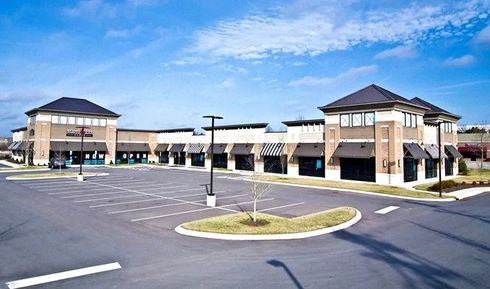 A modern, U-shaped retail strip center with tan brick facades, dark awnings, and an empty parking lot under a blue sky.