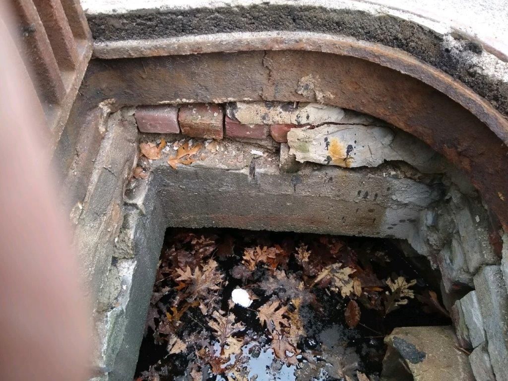 A manhole cover with a hole in it and leaves on the ground.