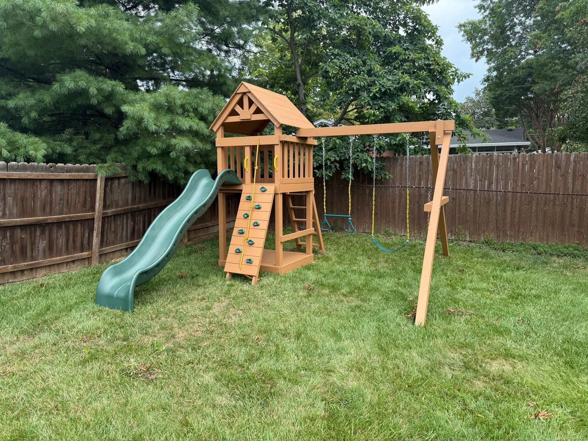 Wooden playground equipment in a yard with swings, slide, climbing wall, and a playhouse.