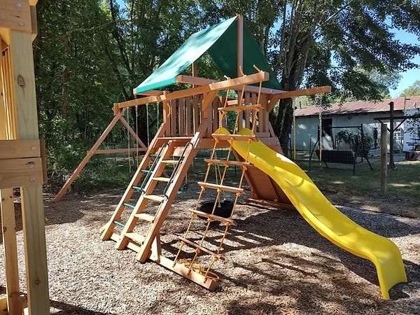 Playground with a green slide, climbing walls, and wooden structure on wood chips, under blue sky.