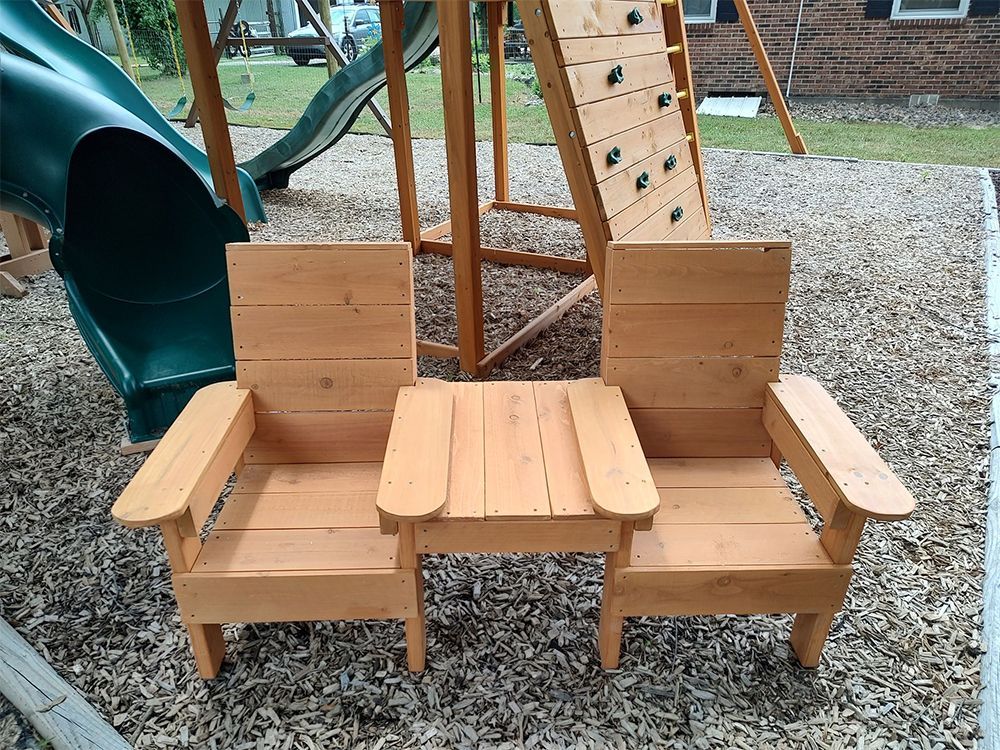 Wooden two-seat outdoor chair with a small table in the center, near a playset slide and climbing wall.