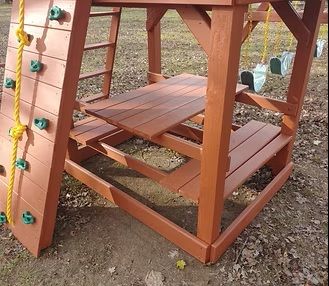 Wooden playset with a table and benches; brown stain; a climbing wall and swings are visible.