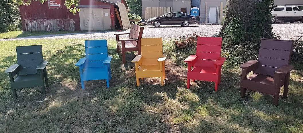 Five colorful Adirondack chairs sit on grass; a barn and a car are in the background.
