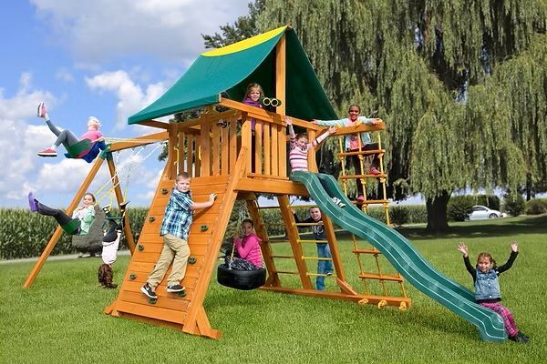 Children playing on a wooden playground set with swings, slide, climbing wall, and rope ladder.