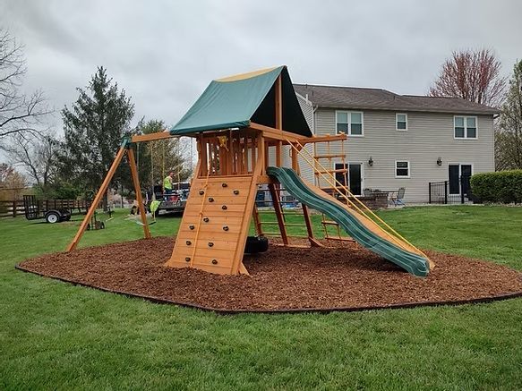 Wooden playset with swing set, rock climbing wall, slide, and wood-chip ground covering in a backyard.