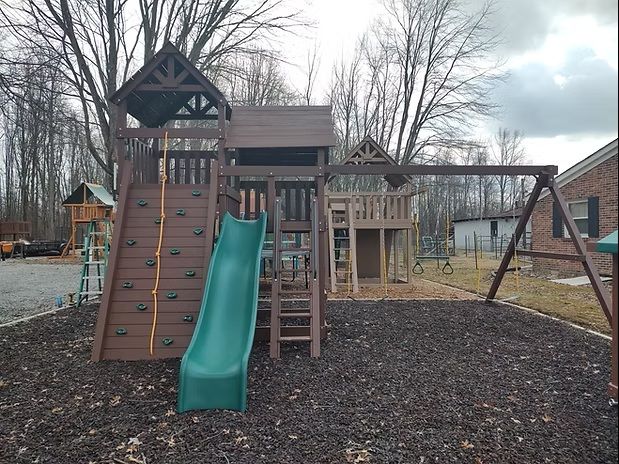 Playground with a slide, climbing wall, swings, and playhouse on a mulched surface.