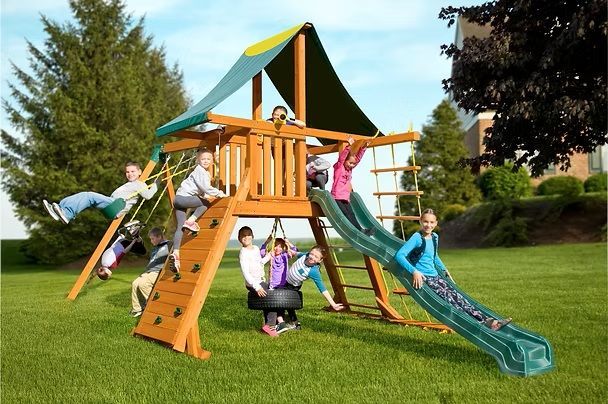 Children playing on a wooden outdoor playset with a slide, climbing wall, swings, and tire swing in a grassy yard.