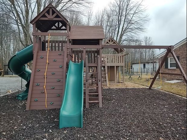 Playground set with a slide, climbing wall, swings, and playhouse in a yard.