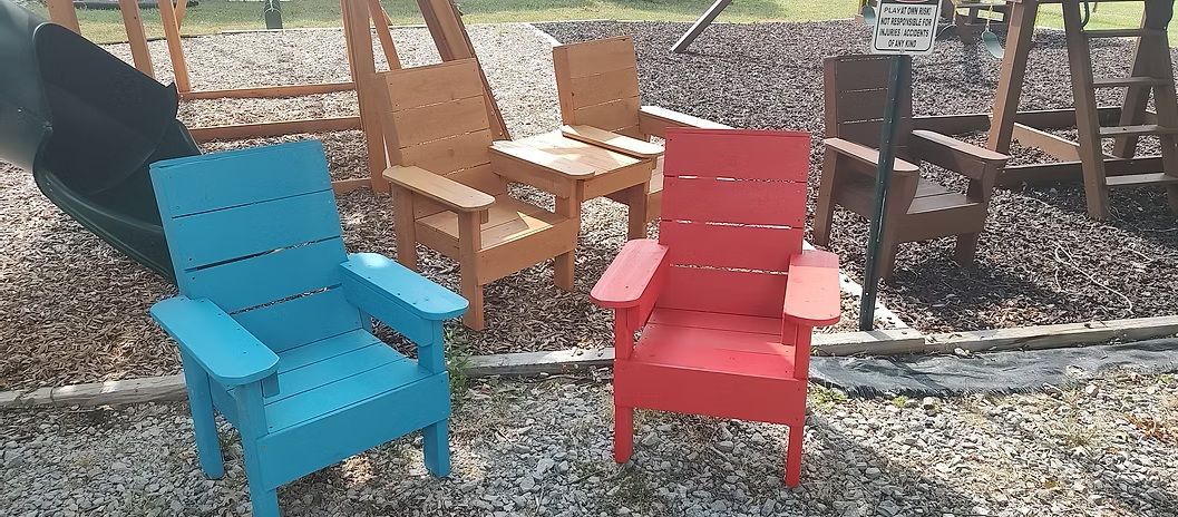Three brightly-colored Adirondack chairs on a pebble surface. A blue chair is in the front, with red and brown chairs behind.