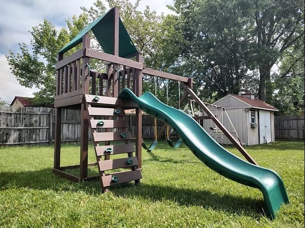 Playground set with green slide, swings, and wooden tower in a grassy backyard.