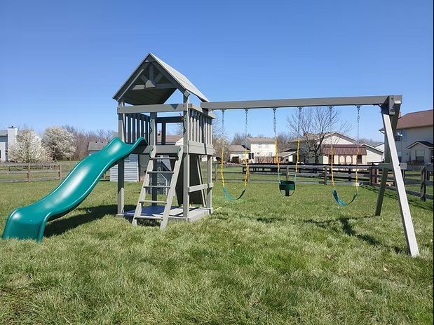 Playground set with a green slide, wooden tower, and swings on a grassy lawn with a blue sky.