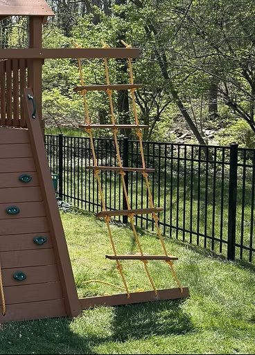 Wooden playset with climbing rope ladder and green lawn. Black fence and trees in background.