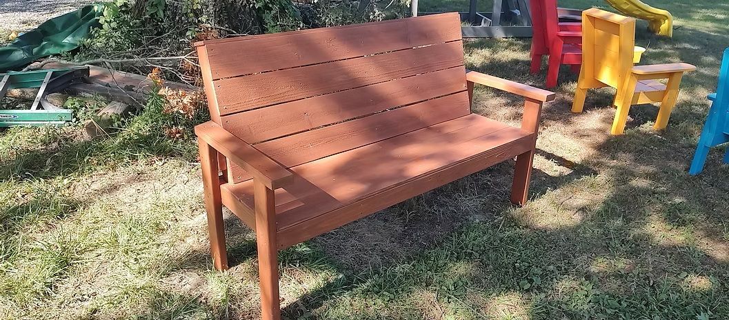 Brown wooden bench on grass with colorful chairs in the background.
