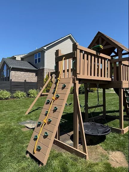 Wooden playground with climbing wall and rope ladder in a grassy backyard, sunny day.