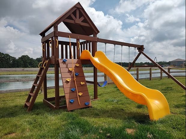 Wooden playground set with climbing wall, slide, and swings on a grassy field.