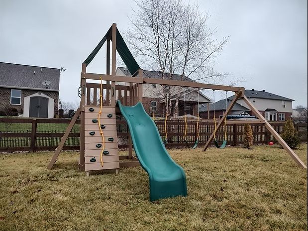 Wooden playground with a slide, climbing wall, and swings in a grassy yard under a cloudy sky.