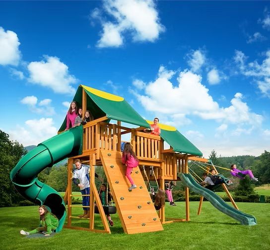 Children playing on wooden playground set with slide, climbing wall, and swings, green grass, blue sky.