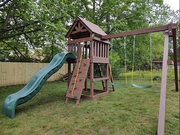 Brown wooden playset with a green slide, climbing wall, and swings in a grassy yard.